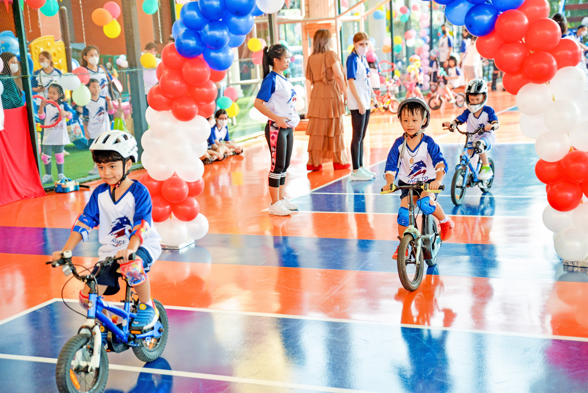 Children riding bicycles through balloon arches on the court